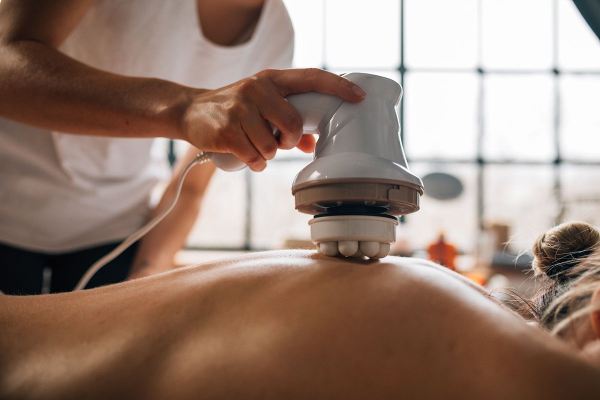A hand guiding a white corded electric massager with rounded nodes across a woman's bare back, with large grid windows blurred behind